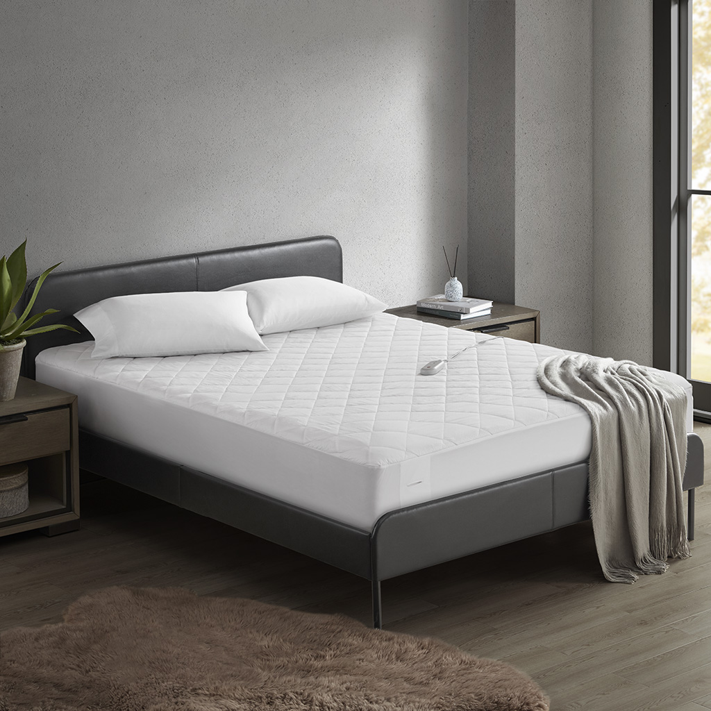 White quilted heated mattress pad fitted on a bed; a single wired hand control is visible